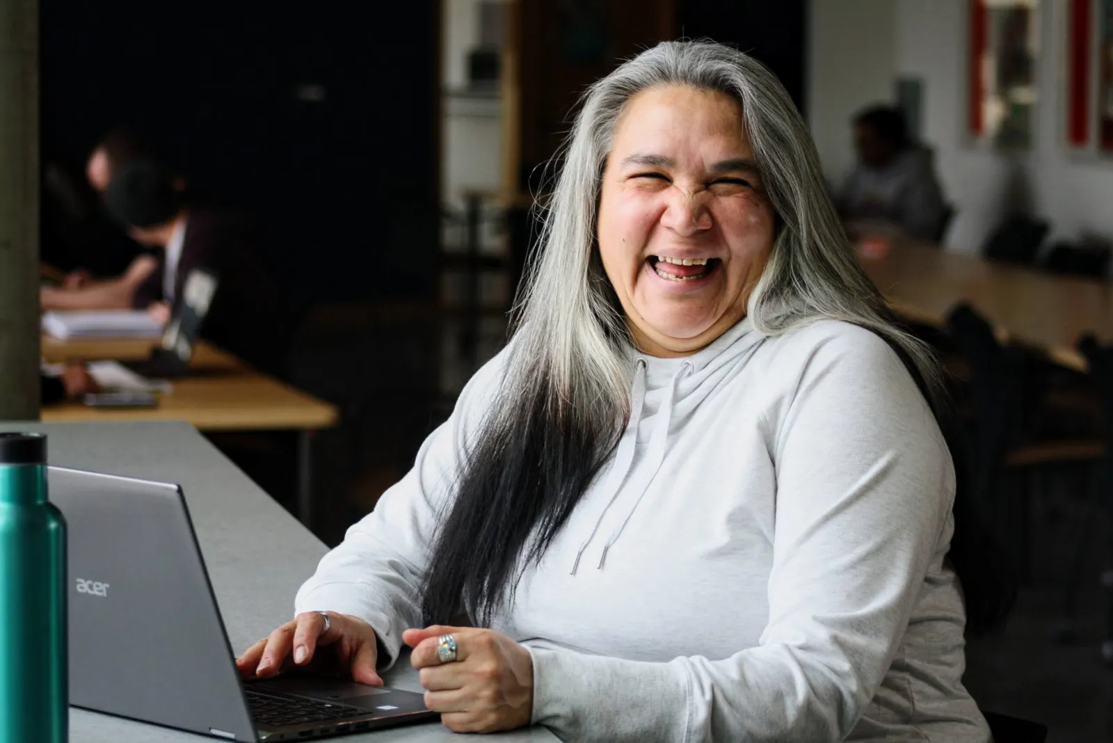 A woman with long grey hair laughing at her laptop in a bright study space