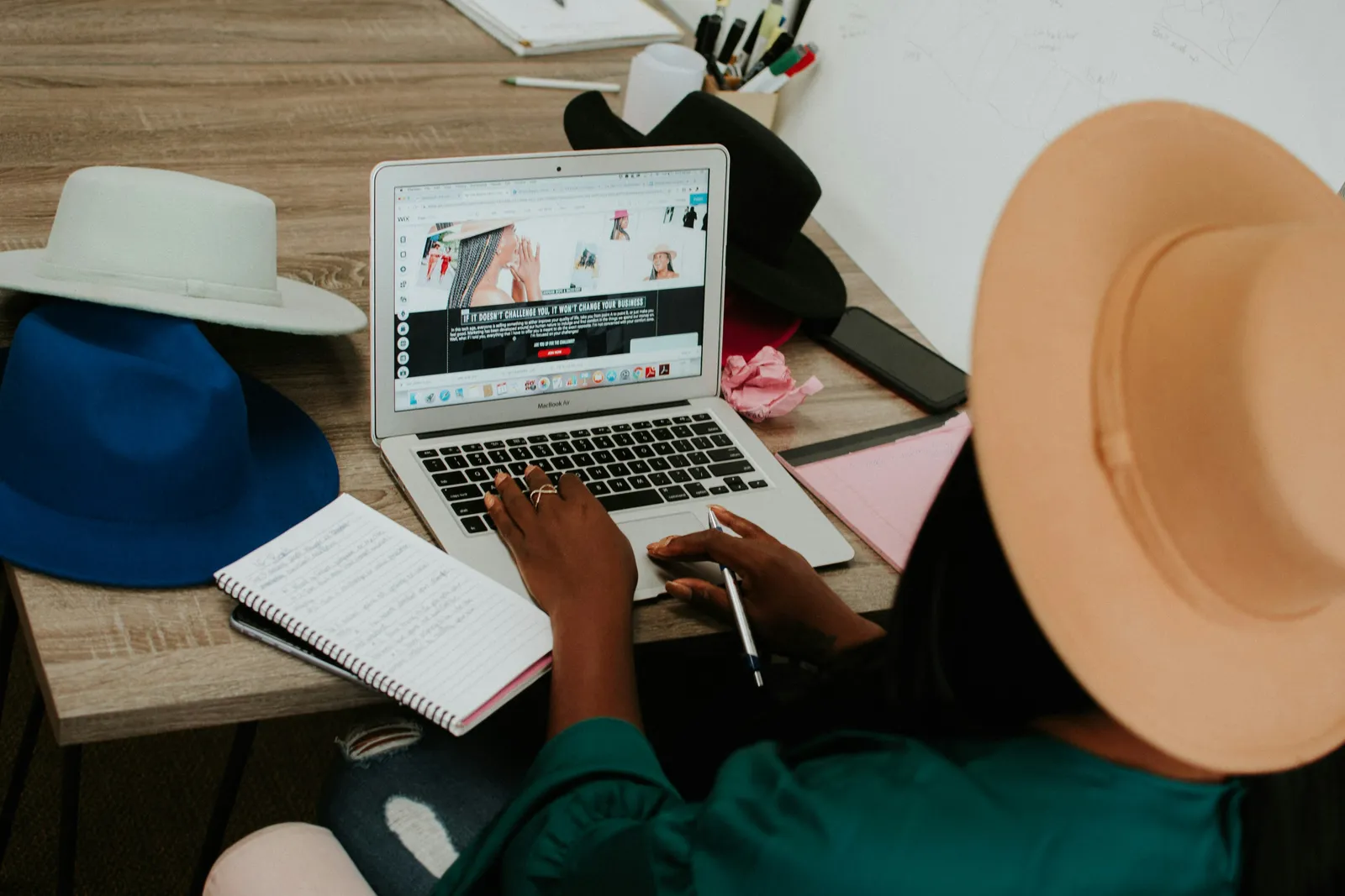 Over the shoulder of a hat maker at her desk, editing her shop’s website with her notes and a phone beside the laptop