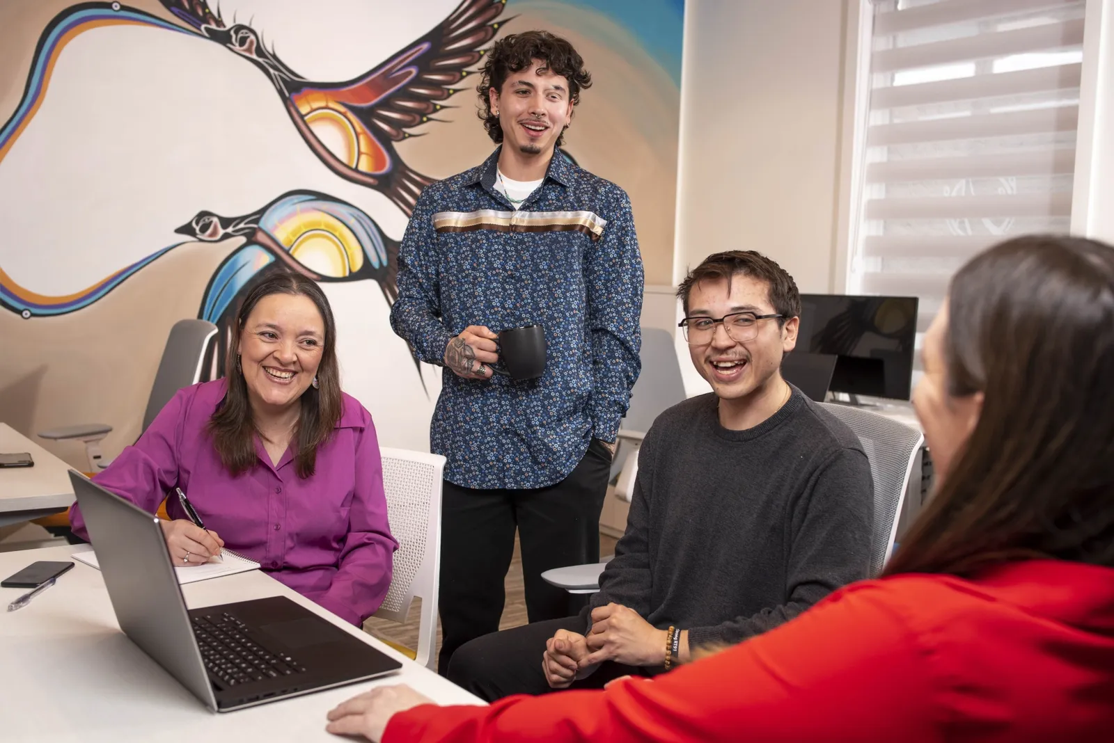 Four people laugh together around a table with a laptop, one standing with a mug, a large Indigenous mural of birds on the wall behind them