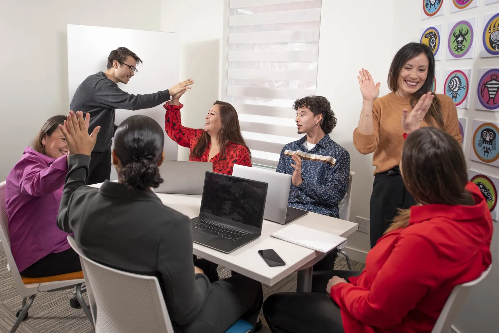 Coworkers trading high fives and applause around a meeting table of laptops, with Indigenous art on the wall behind them