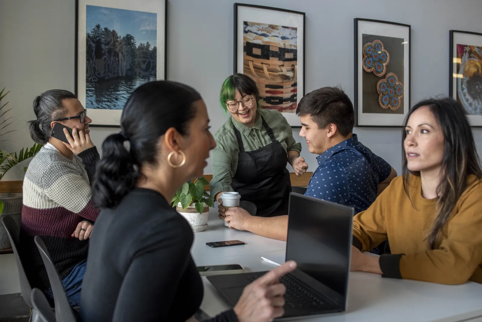 A mixed group around a shared table, one on the phone, one in an apron, and laptops open between them