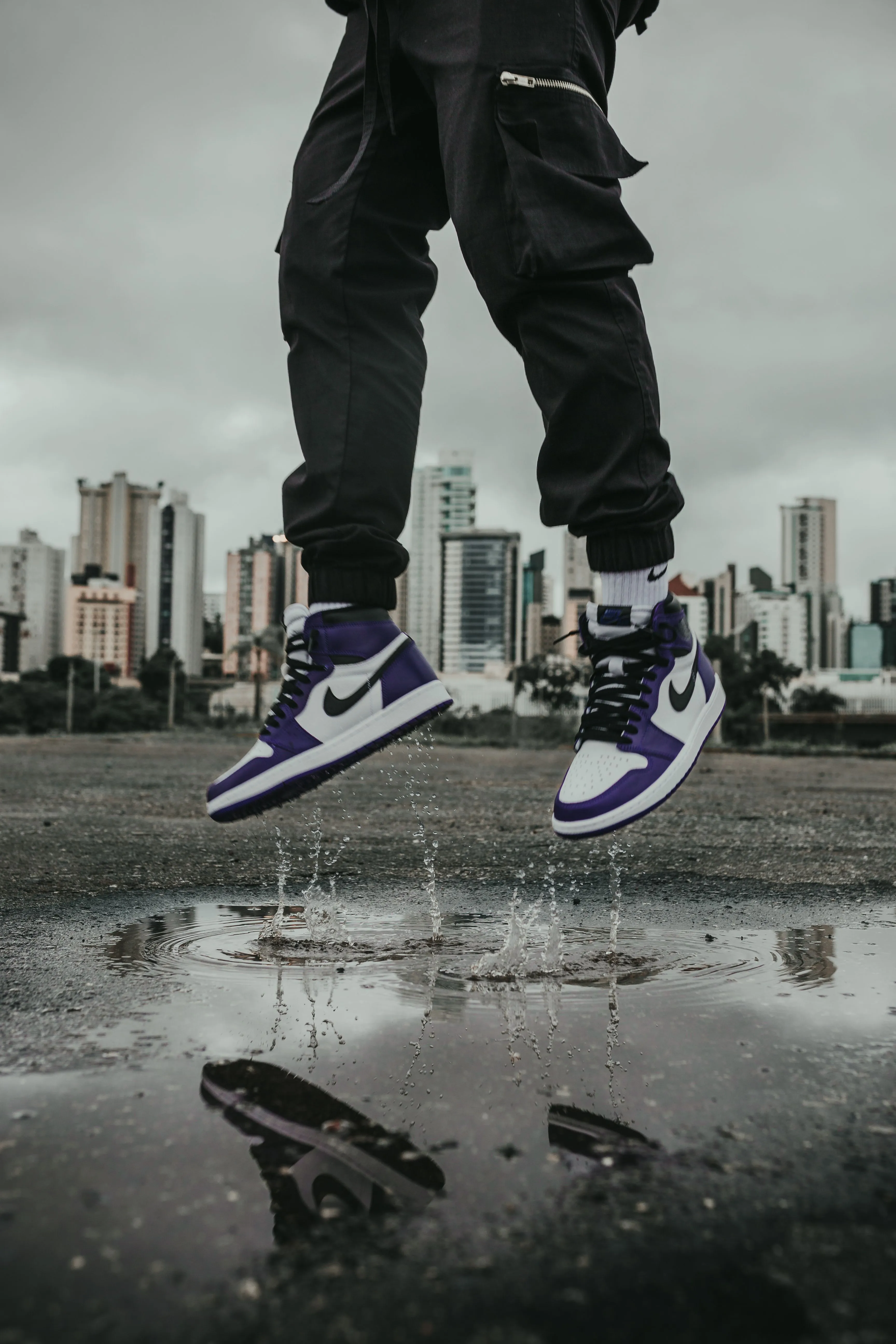 A person in high-top sneakers jumping over a rain puddle with a city skyline behind them