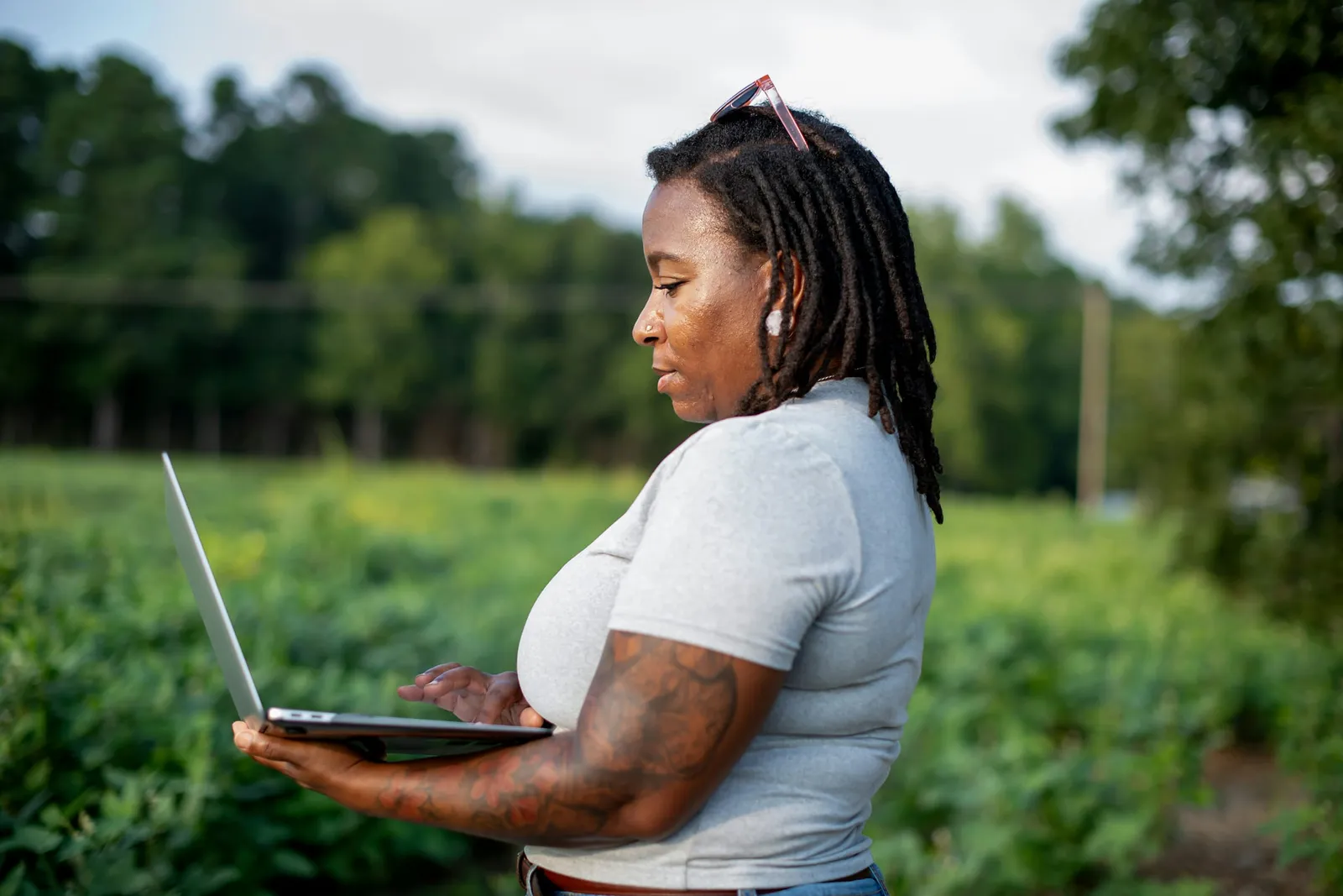 A farmer standing in her field, typing on a laptop balanced on one arm