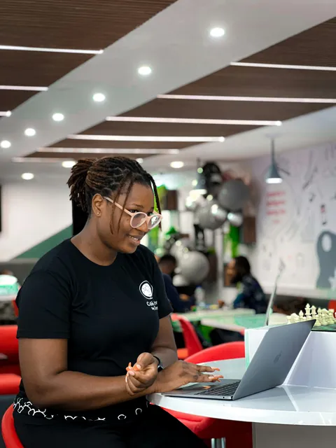 A young woman smiling as she types on her laptop in a busy café