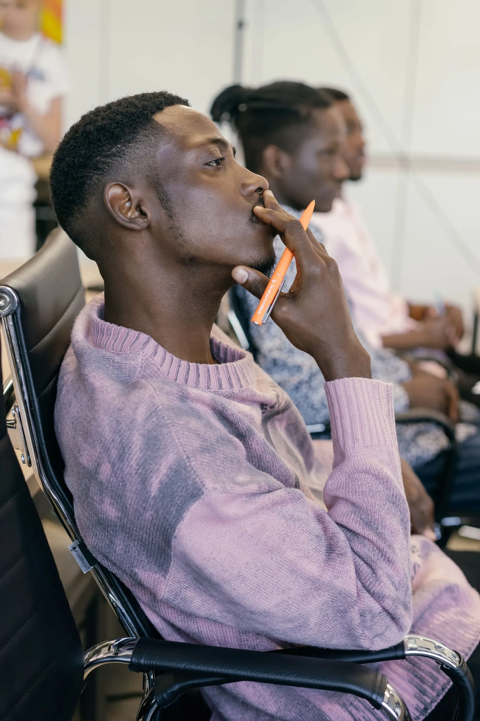 A young man in a lilac sweater listening closely in a workshop, a pen at his lips, others seated along the row
