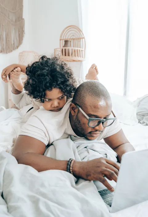 A father on a bed with his laptop, his small child draped over his shoulder watching the screen