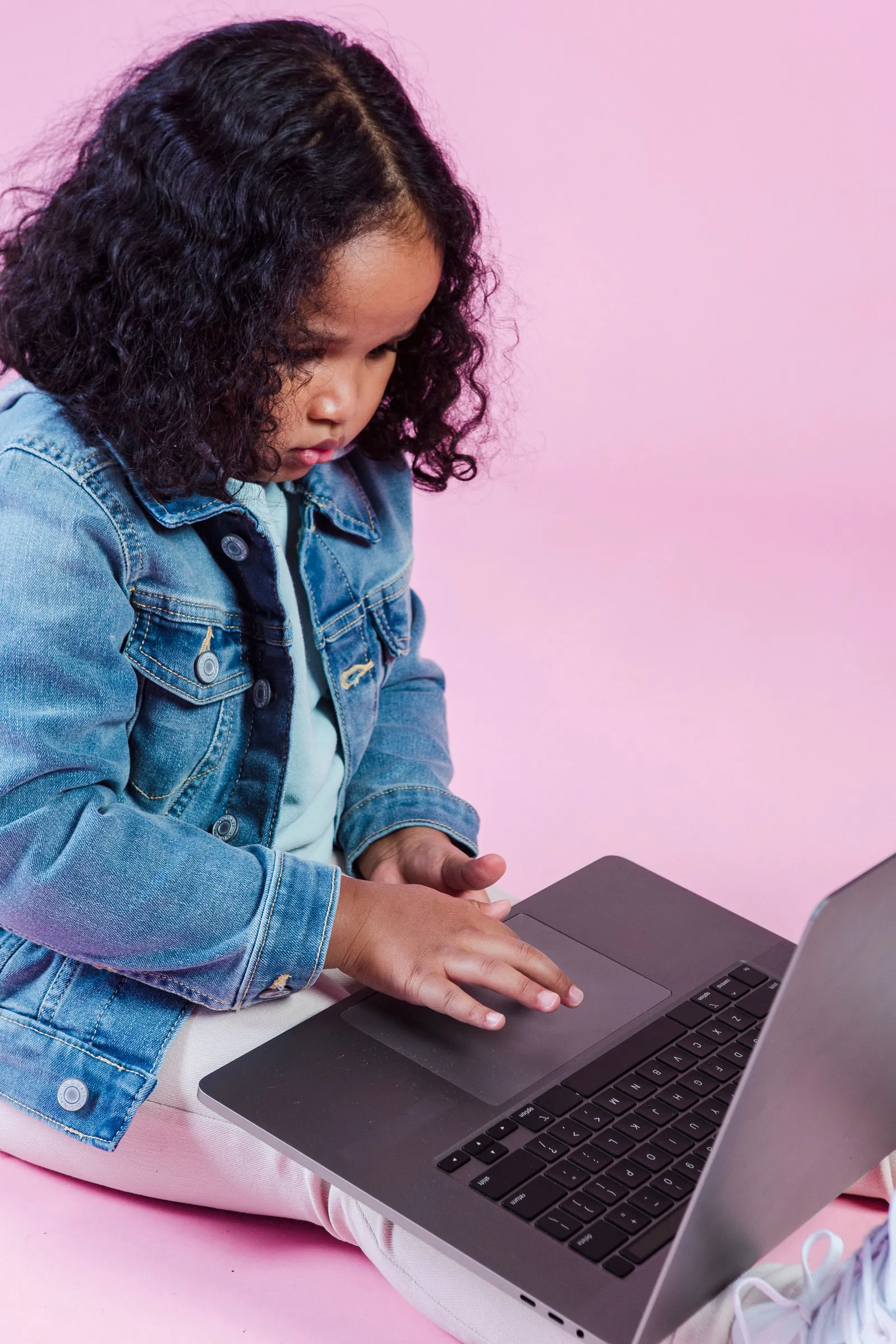 A young girl in a denim jacket concentrating on a laptop in her lap, against a pink backdrop