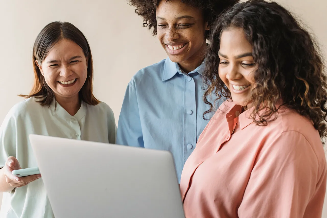 Three women standing shoulder to shoulder, laughing at something on a laptop