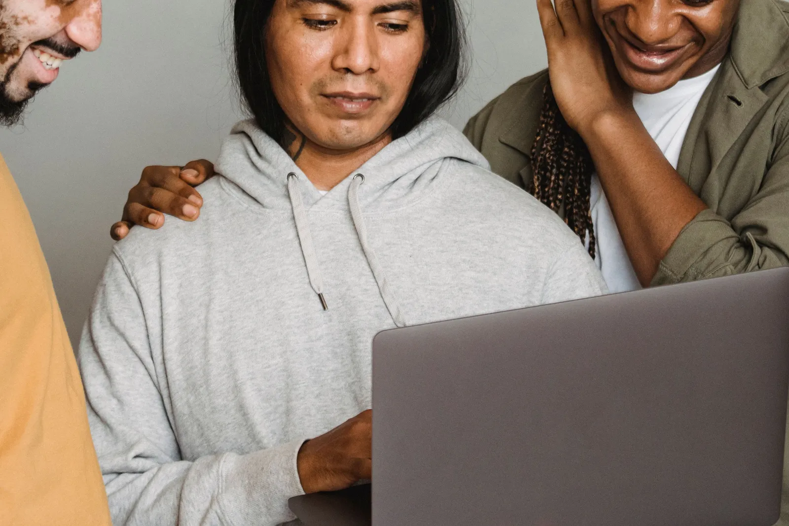 Three people gather around a laptop, one resting her hand on the shoulder of the man at the keyboard as she laughs