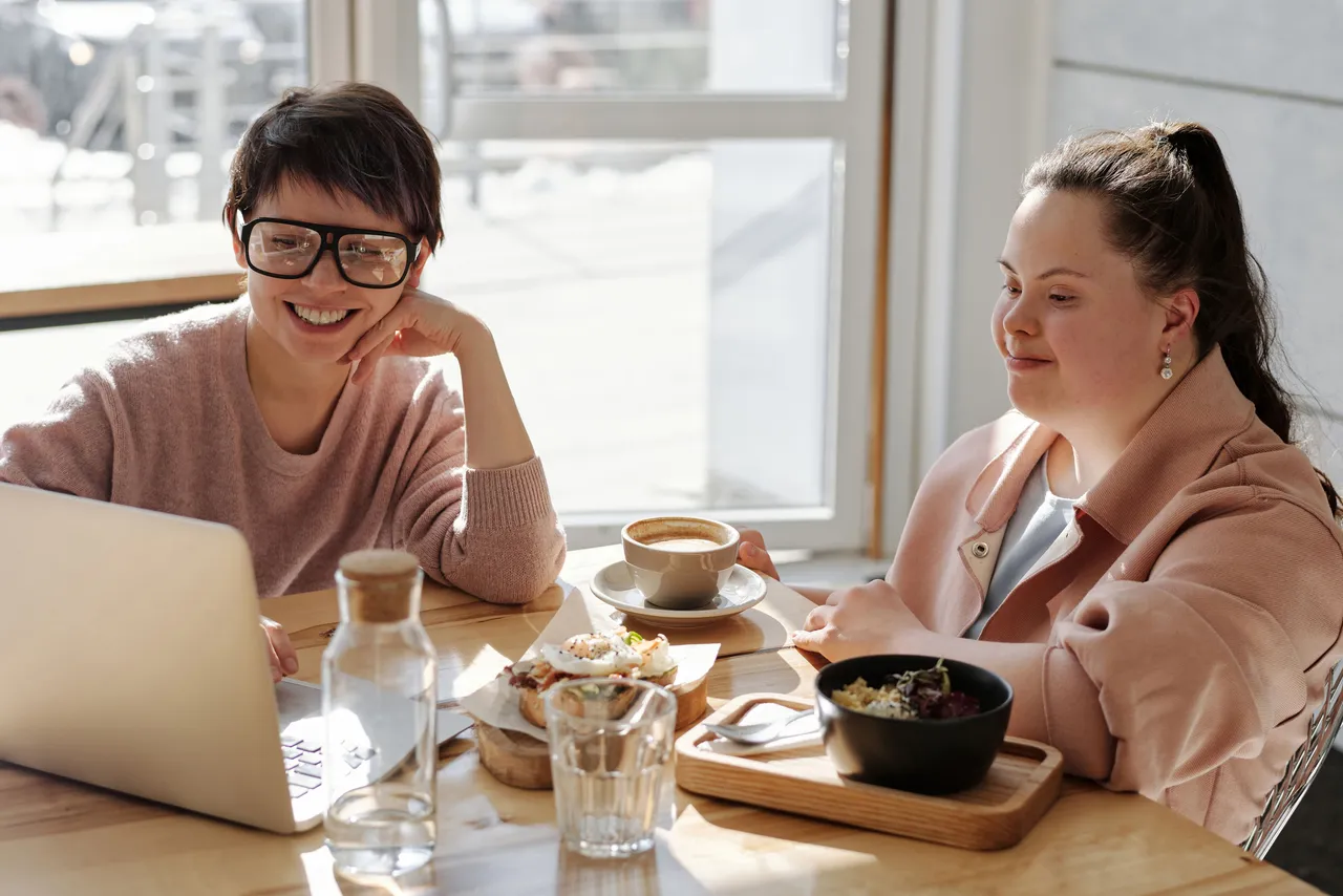 Two women smiling at a laptop together at a sunny cafe table