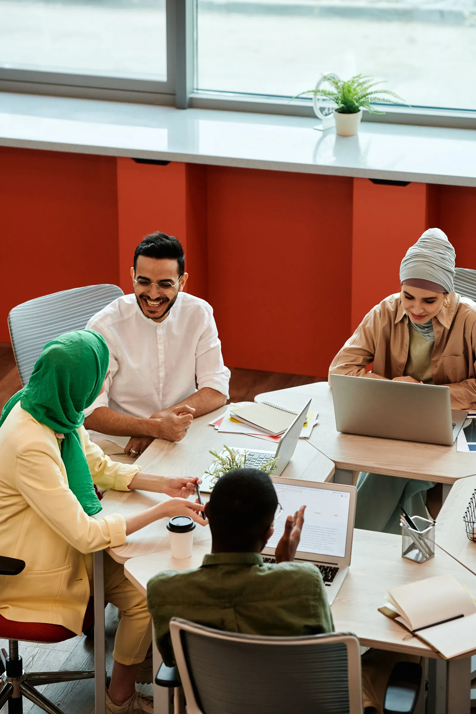 Four colleagues laugh together around shared desks with laptops and notebooks, two of them wearing hijabs