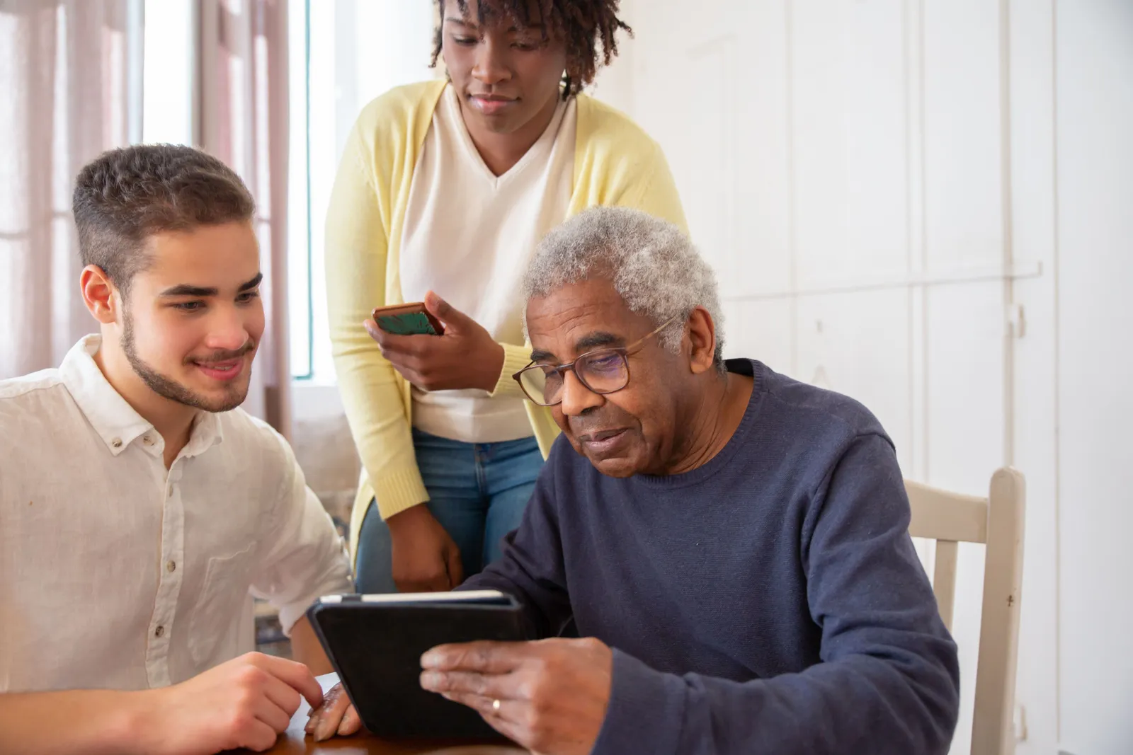 Three generations around a kitchen table, an older man reading a tablet while a younger man and a woman look on