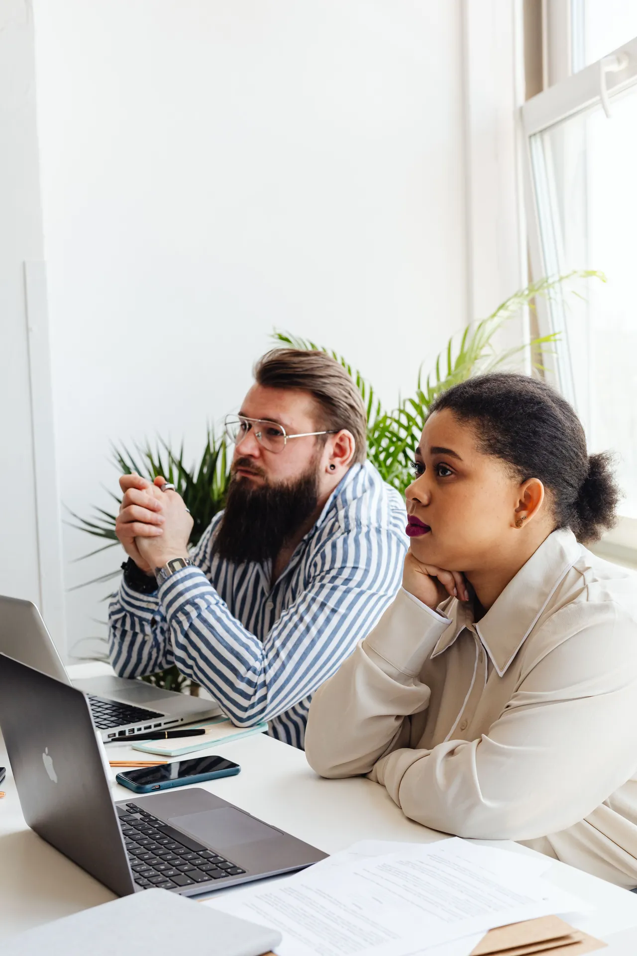 Two colleagues at a bright desk with laptops and papers, listening to someone off camera