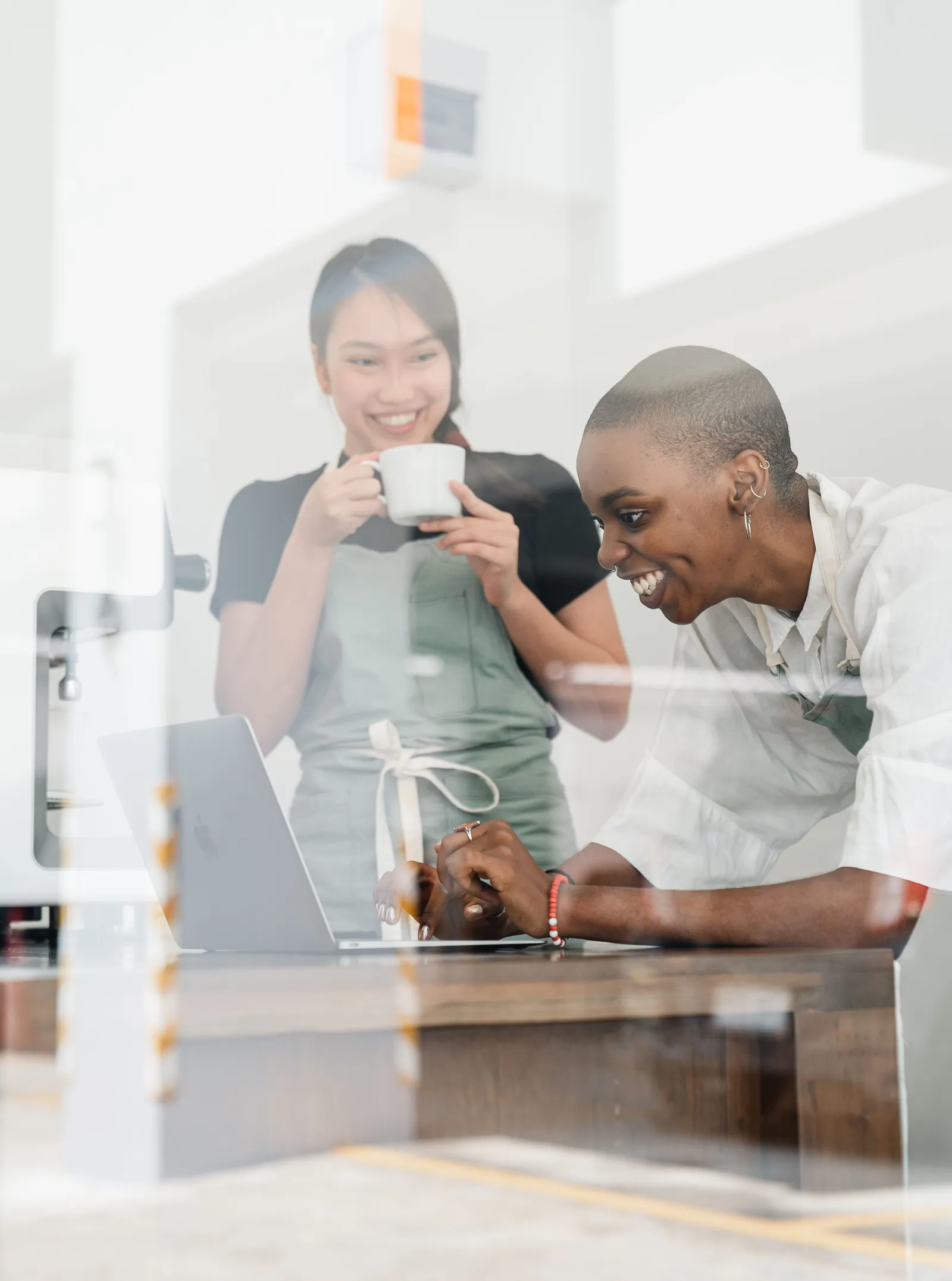 Two cafe workers in aprons laughing at a laptop on the counter, seen through the window