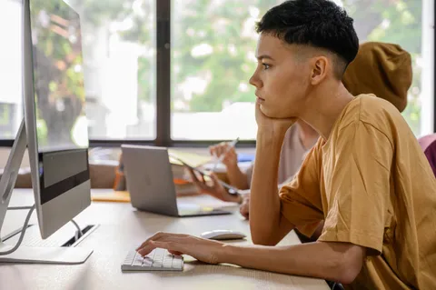 A young person working at a computer at a shared table, with other learners behind