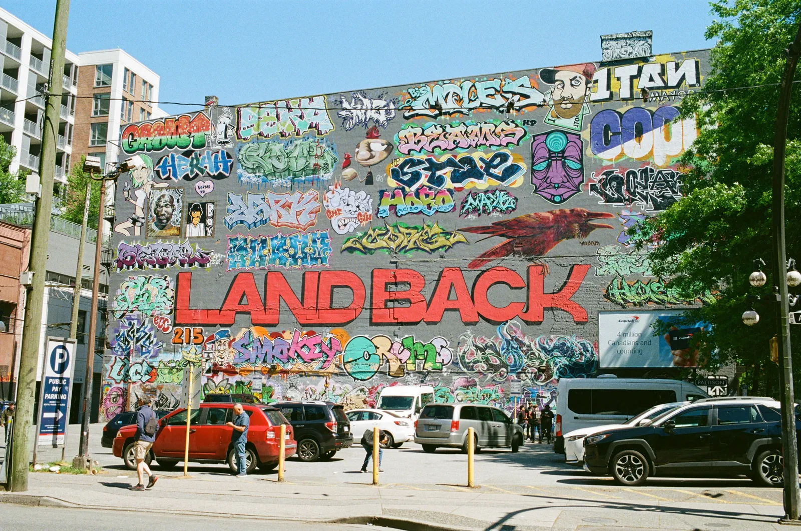 A LAND BACK mural painted in red across a graffiti-covered wall, cars parked along the street in front of it
