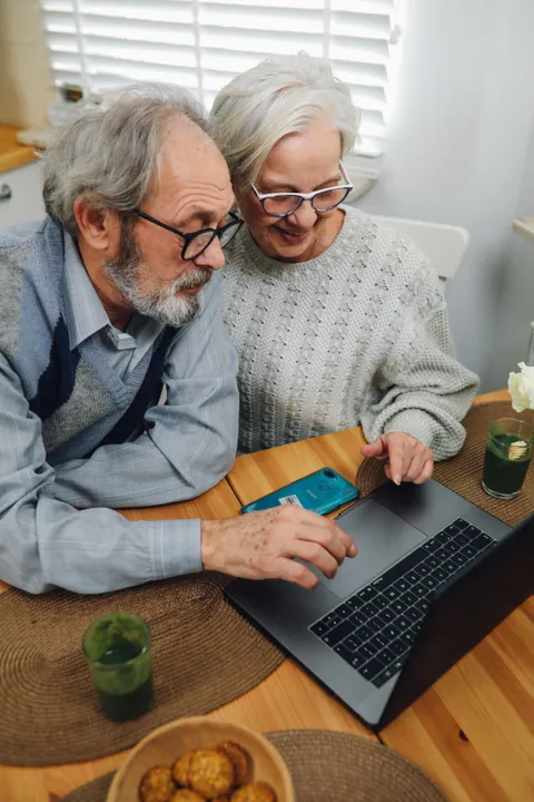 An older couple side by side at a laptop on their kitchen table