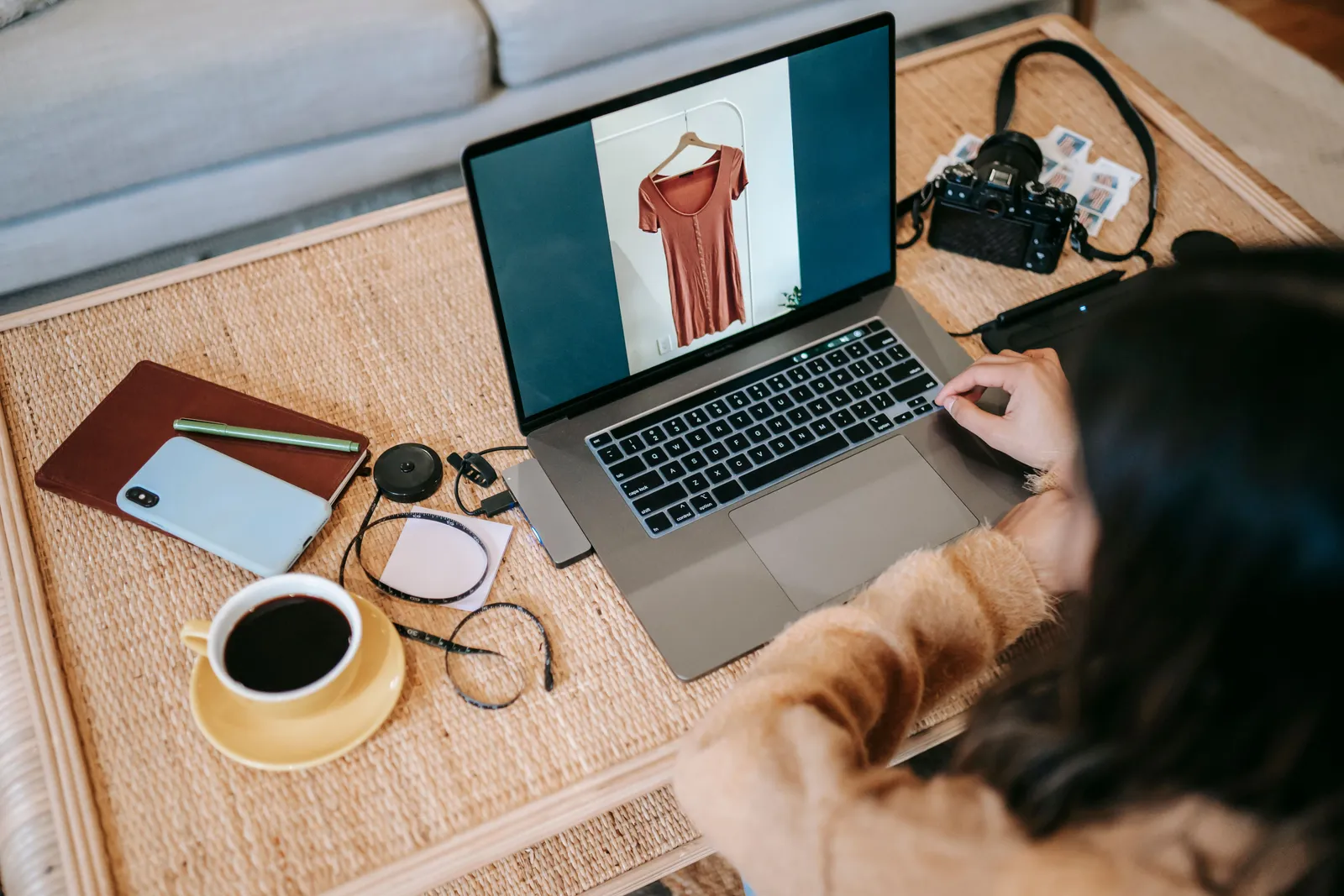 Someone at a laptop on a woven coffee table, a shop page open beside a camera, a notebook, and a cup of coffee
