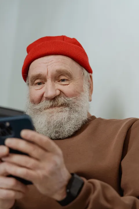 An older man with a white beard and a red knit beanie smiling at the phone in his hands