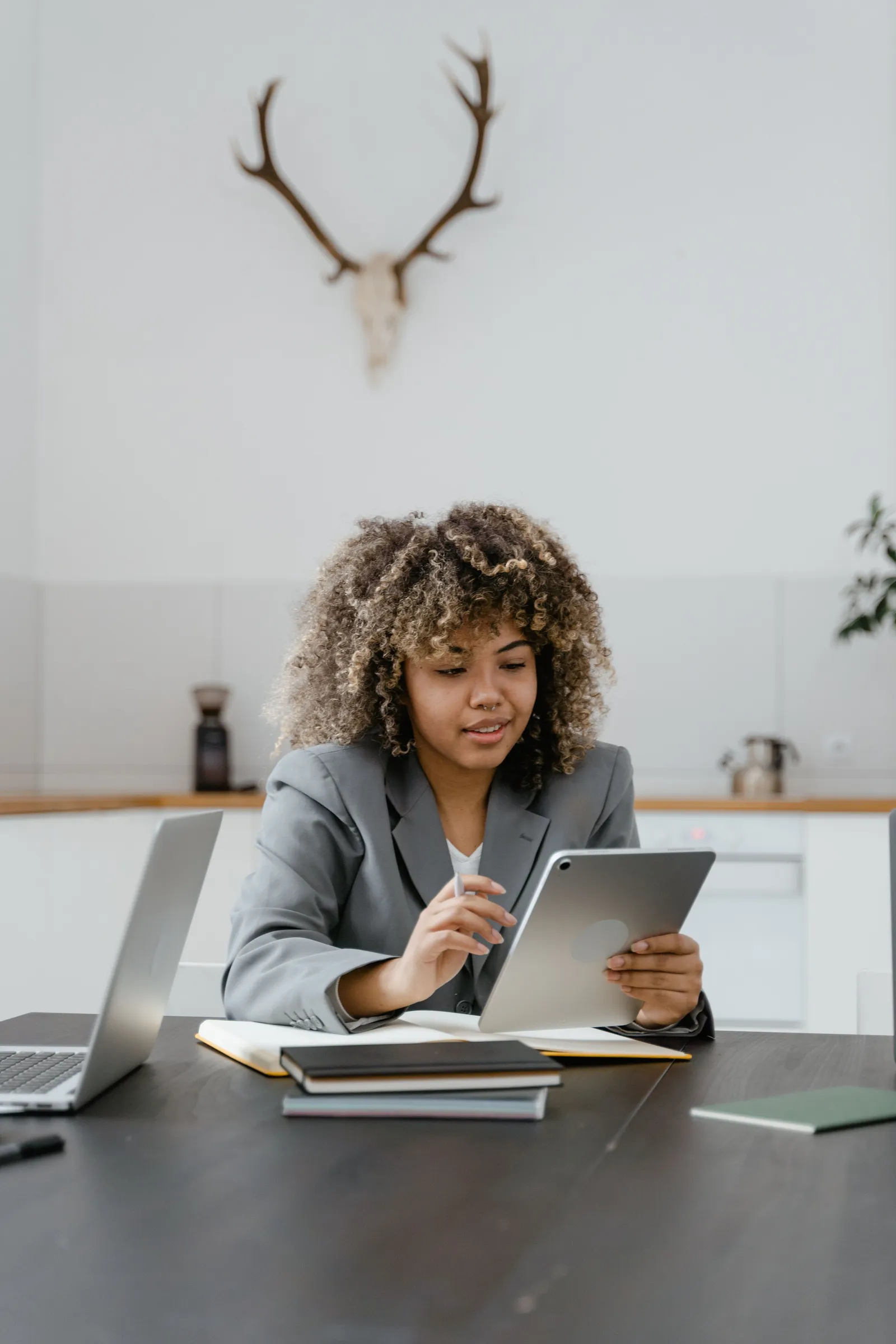 A woman in a blazer working from a tablet at a desk, a laptop and notebooks open beside her