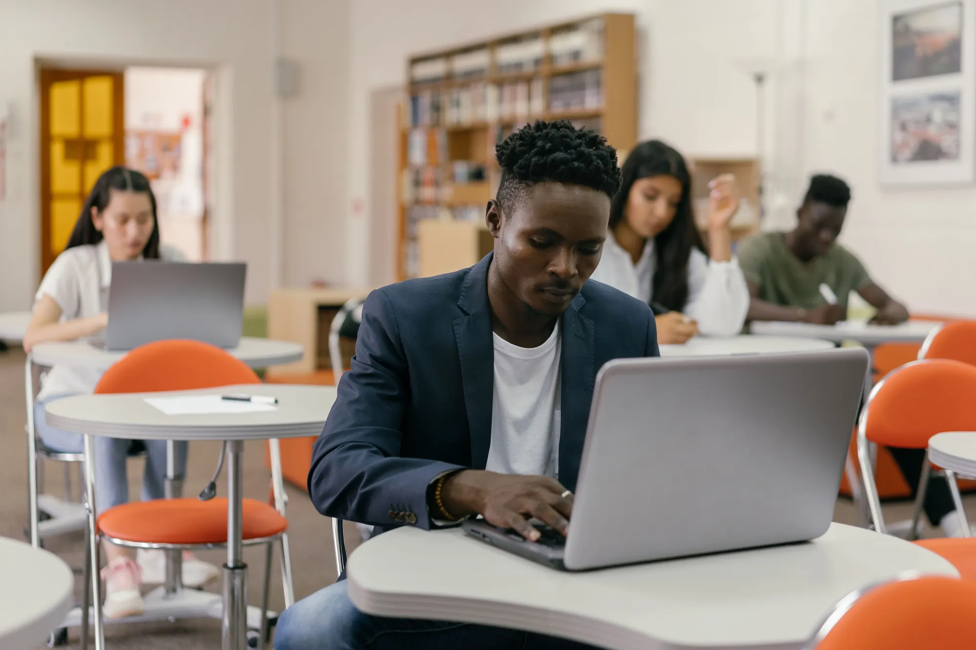 A man in a blazer typing on a laptop in a bright classroom, other learners at the tables behind him
