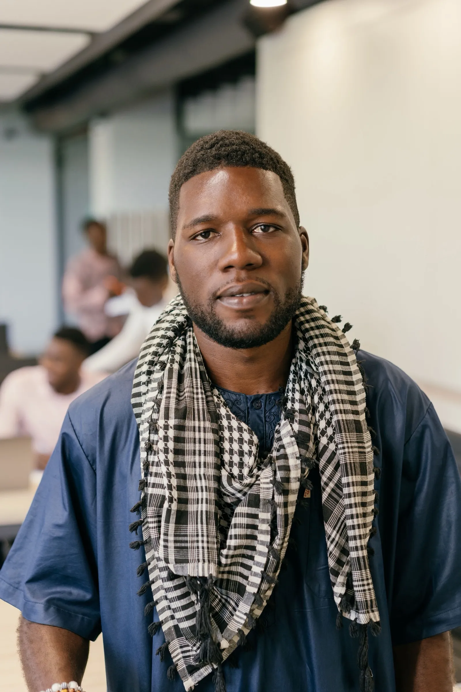 A young man in a blue shirt and a checked scarf standing in a classroom, other learners at the tables behind him