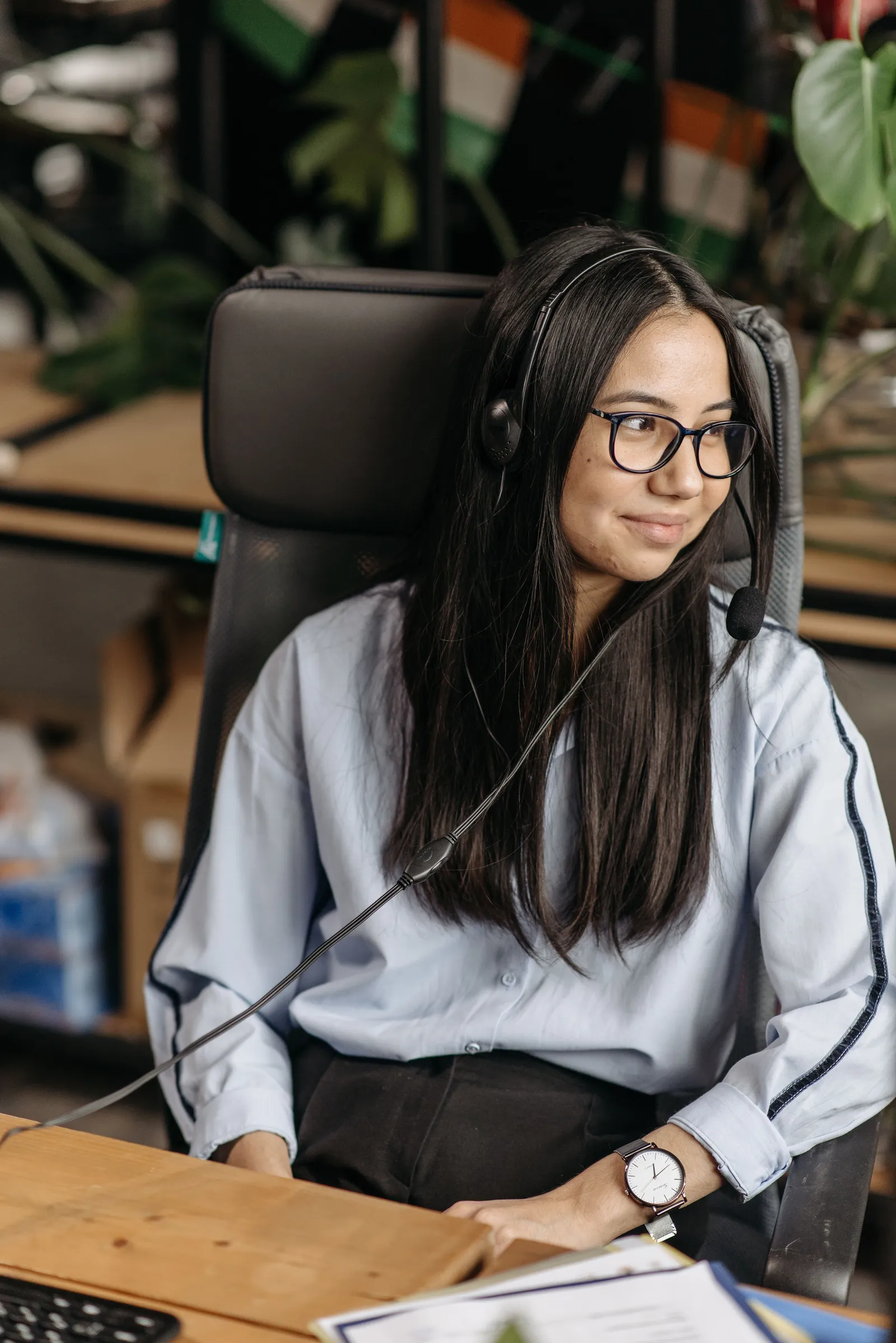 A young woman in glasses and a headset at her desk, smiling as she looks across the office