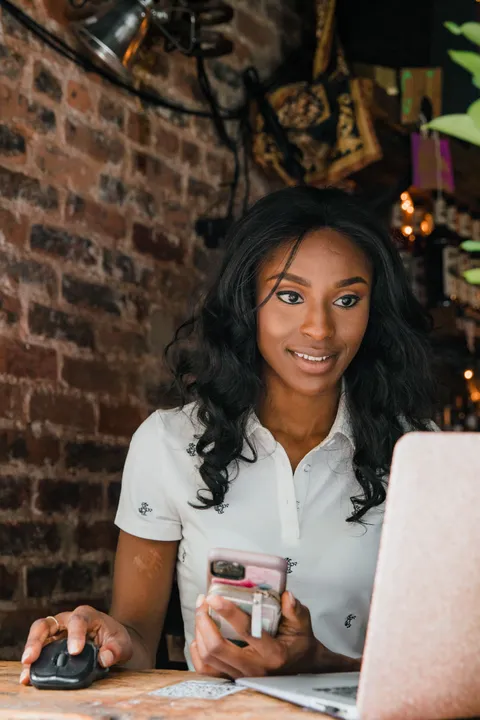 A woman working at a laptop, phone in hand, at a wooden table in a brick-walled cafe