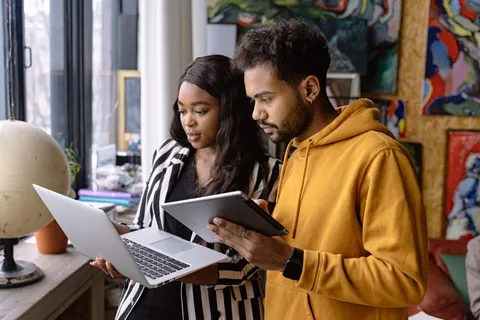 Two young artists looking at a laptop and a tablet together in a studio hung with colourful paintings