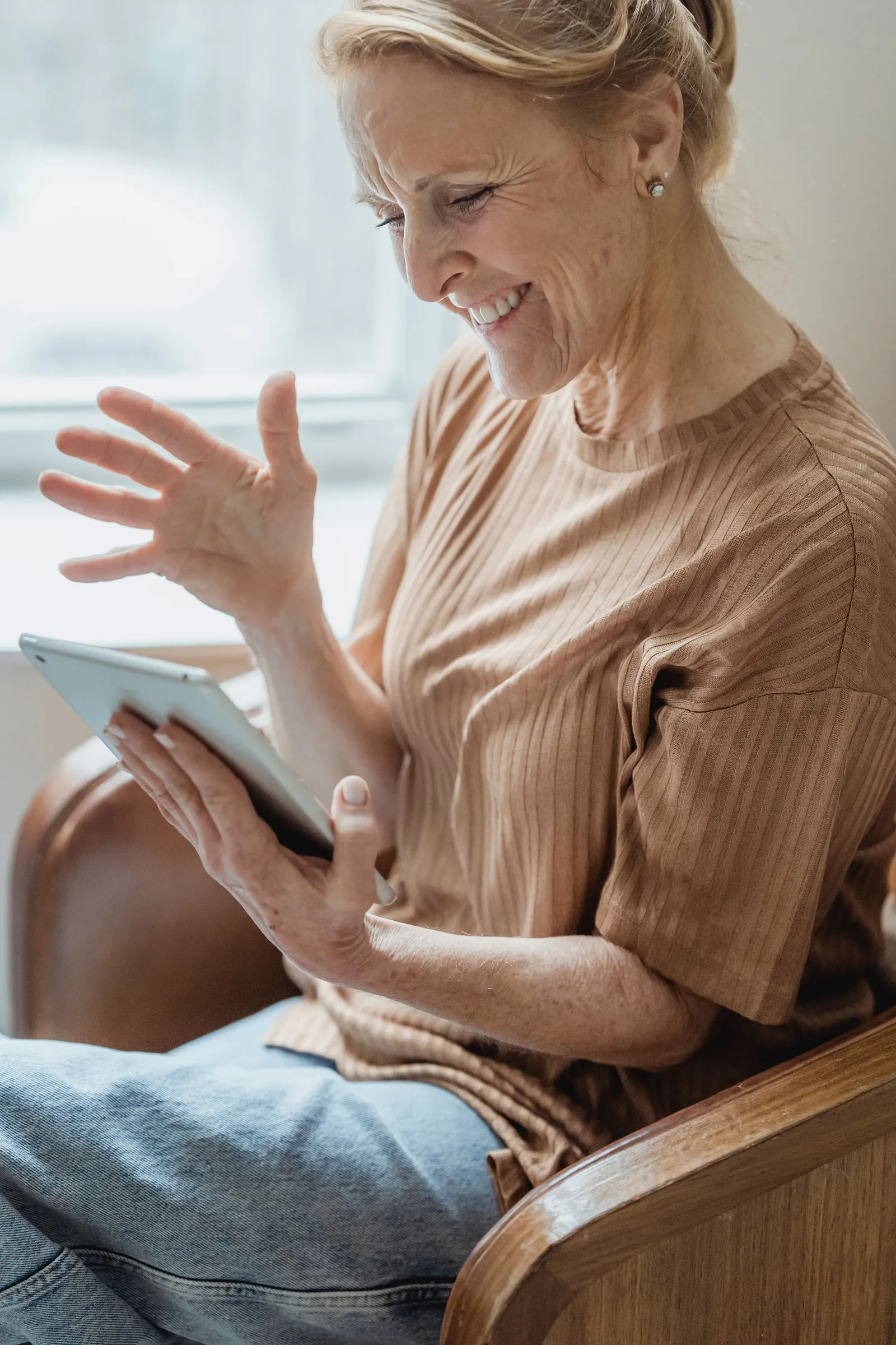 An older woman in an armchair smiling and waving at a video call on her tablet