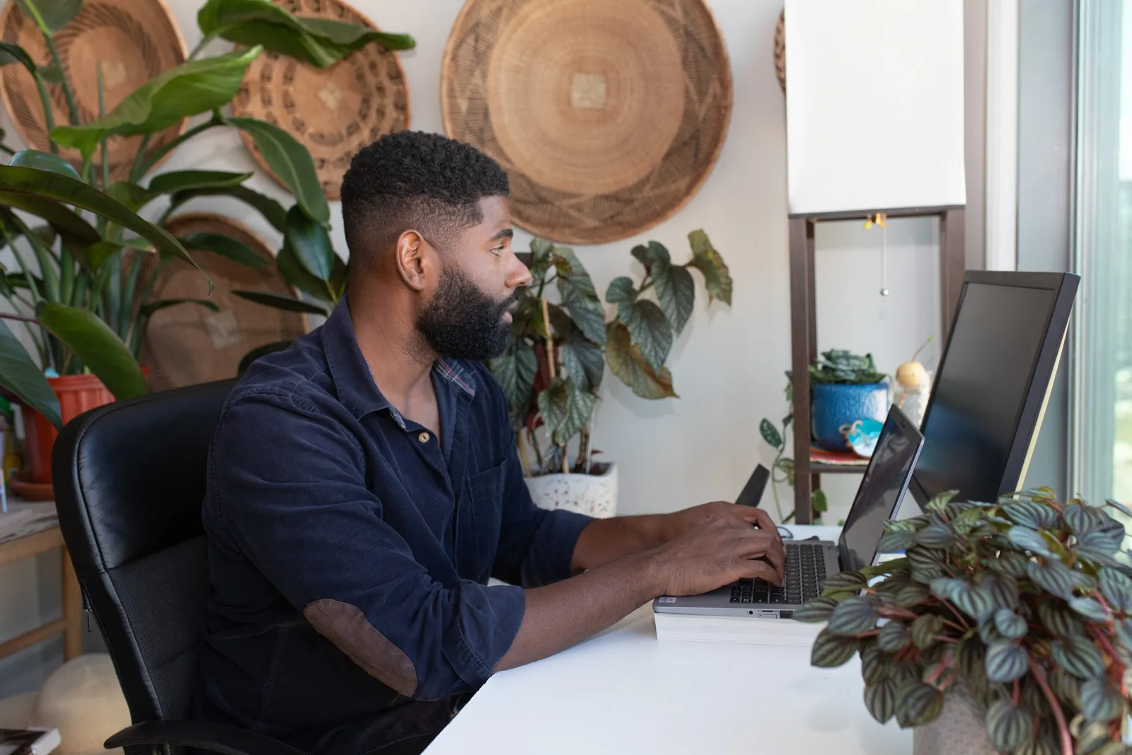 A man typing at a laptop with a second monitor in a bright home office full of plants