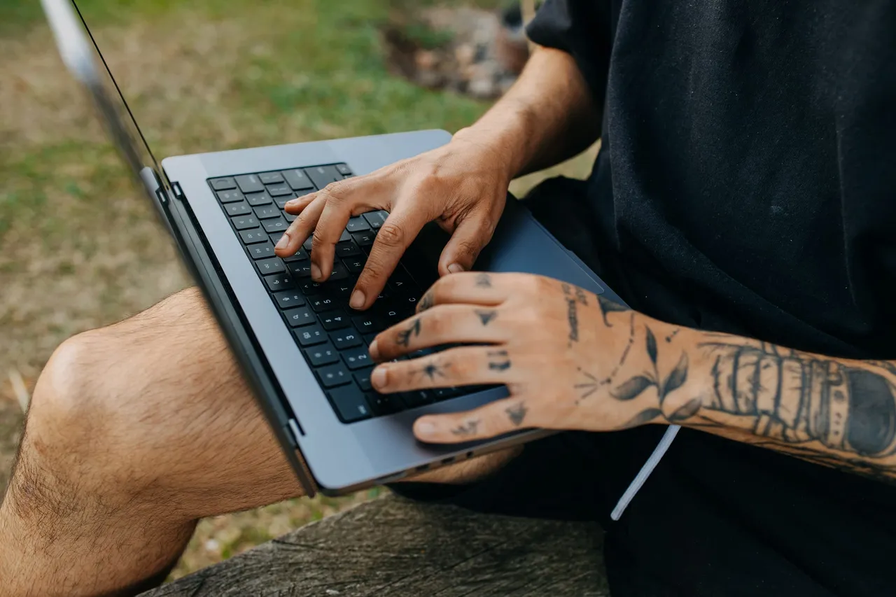 Tattooed hands typing on a laptop balanced on someone’s knees, outdoors on the grass