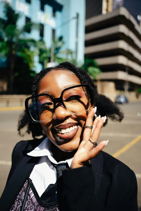 A young Black woman in square glasses and a black blazer laughing, one hand to her cheek
