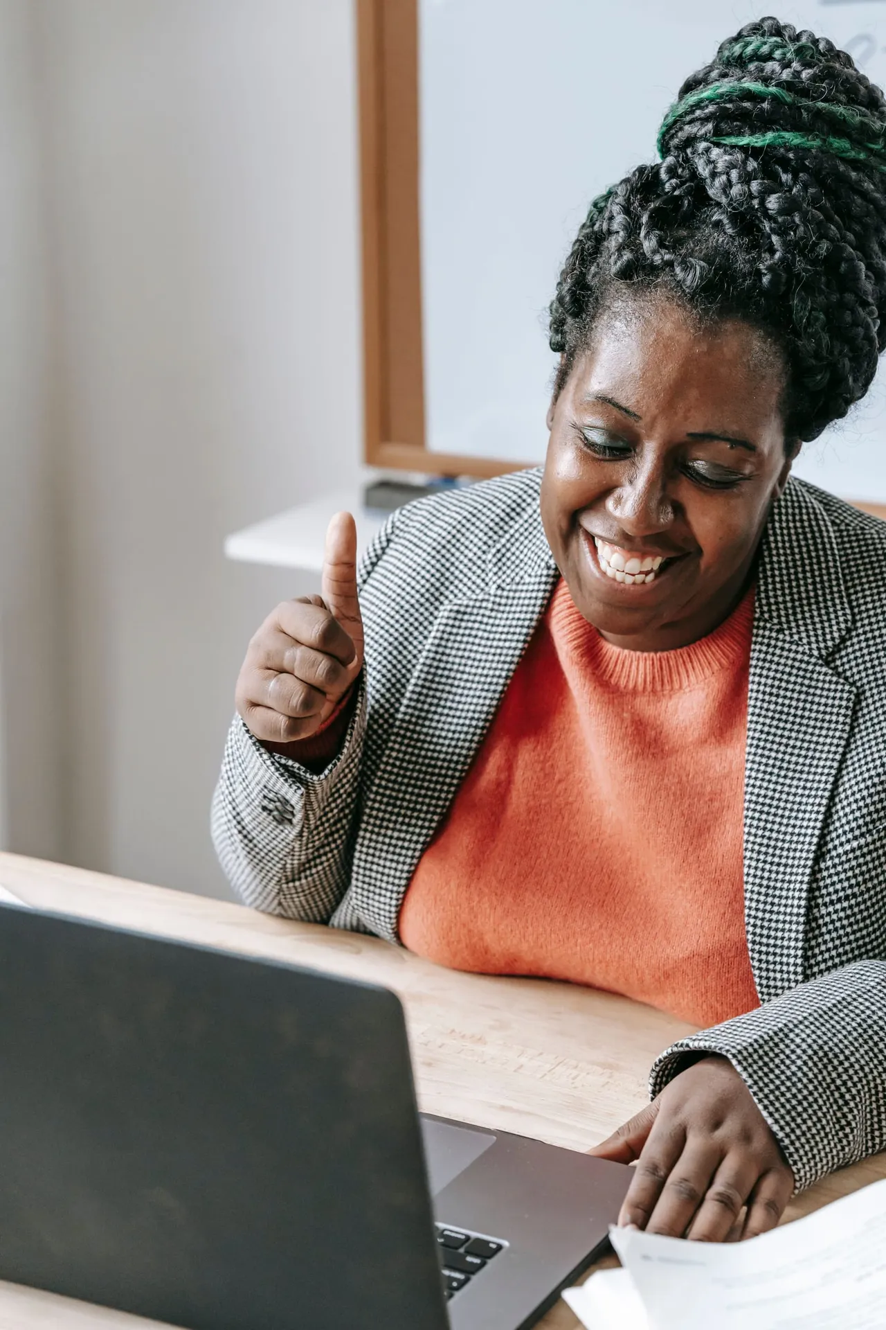 A woman in a houndstooth blazer smiling and giving a thumbs up at her laptop, one hand on printed papers